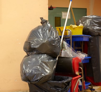 The image depicts a cluttered indoor or outdoor storage area containing several large black refuse bags stacked on top of each other, with some leaning against a yellow-colored wall. The bags appear to be filled with waste materials and are made of durable plastic with a shiny surface. To the right of the bags, there is a red wheeled trolley or bin, partially visible, likely used for transporting rubbish. In the background, there is a blue shelving unit or cart holding a yellow plastic bin, possibly for small waste collection or segregation. The environment looks like a designated rubbish storage space for a commercial or residential property, with an industrial or utilitarian atmosphere. The overall scene emphasizes waste collection and disposal, consistent with the services offered by Commercial Waste Reading in the Reading area, close to postcodes within that locality. The lighting is neutral and sufficient for detailed visibility of the items, highlighting the textures and colors of the waste containers and associated equipment.