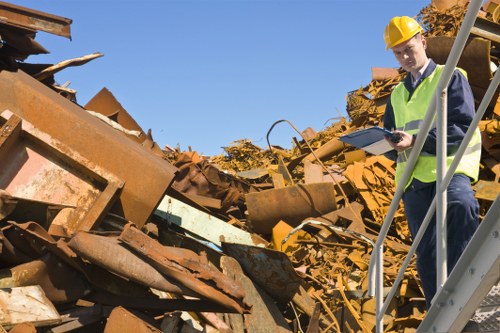 A male worker wearing a yellow safety helmet and a high-visibility vest is standing on a metal staircase at a waste disposal site, holding a clipboard and inspecting a large pile of discarded metal debris. The debris consists of rusted, corroded sheets, beams, and various scrap materials with rough, weathered textures and varying shades of brown, orange, and yellow rust. The background features clear blue sky and parts of the waste site, which appears to be an outdoor industrial area with scattered waste and scrap metal. The scene is well-lit by natural daylight, emphasizing the contrast between the rusted materials and the safety gear of the worker, and reflects the typical environment managed by companies like Commercial Waste Reading specializing in rubbish removal services in Reading.