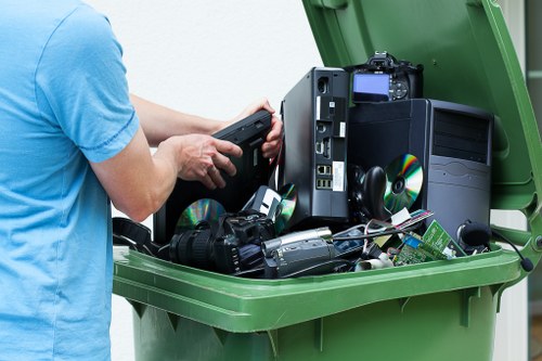 A person wearing a light blue T-shirt is seen pulling a black electronic waste device out of a large green recycling bin. Inside the bin, there are various discarded electronic items, including a desktop computer tower, keyboards, mice, and CDs, all appearing to be in good condition with plastic casings and metallic components. The scene takes place outdoors on a paved surface, and the electronic waste is stacked and visibly visible, indicating collection for disposal or recycling. The image highlights the importance of proper electronic waste disposal services provided by companies like Commercial Waste Reading near Reading, Berkshire, emphasizing responsible rubbish removal for electronic items as part of waste management in the local area.