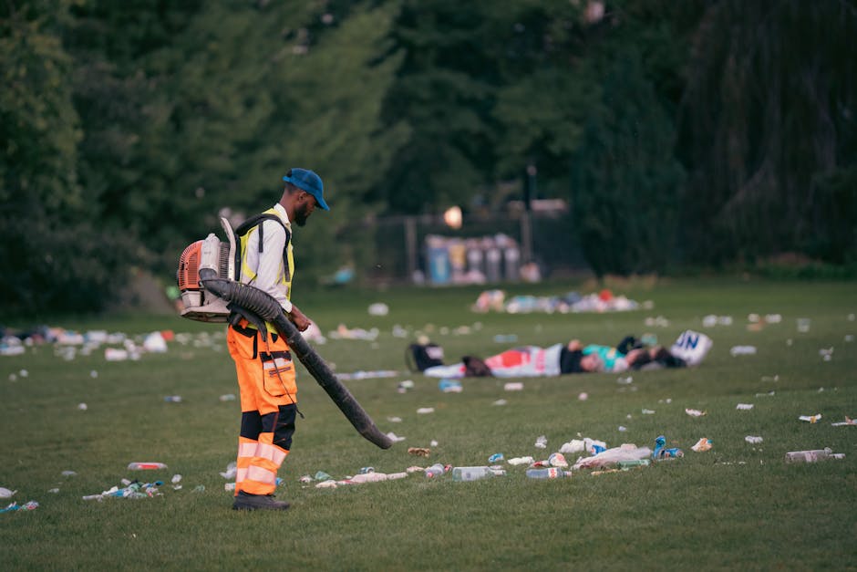 A personnel wearing high-visibility orange trousers, a yellow high-visibility vest, and a blue cap is using a leaf blower with a black hose attachment to clear litter scattered across a grassy area. T