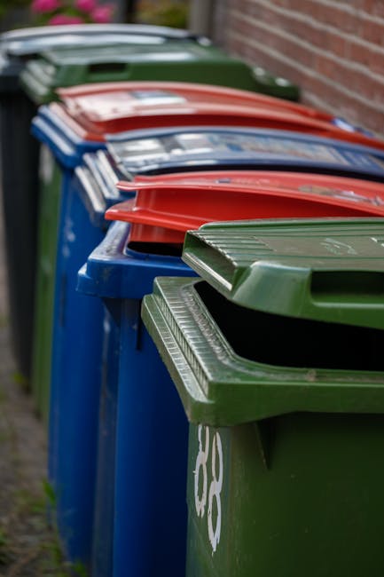 A large outdoor area showing multiple stacks of empty cardboard fruit boxes, predominantly red and yellow with green and white branding, arranged on concrete ground. To the right, there are wire mesh 