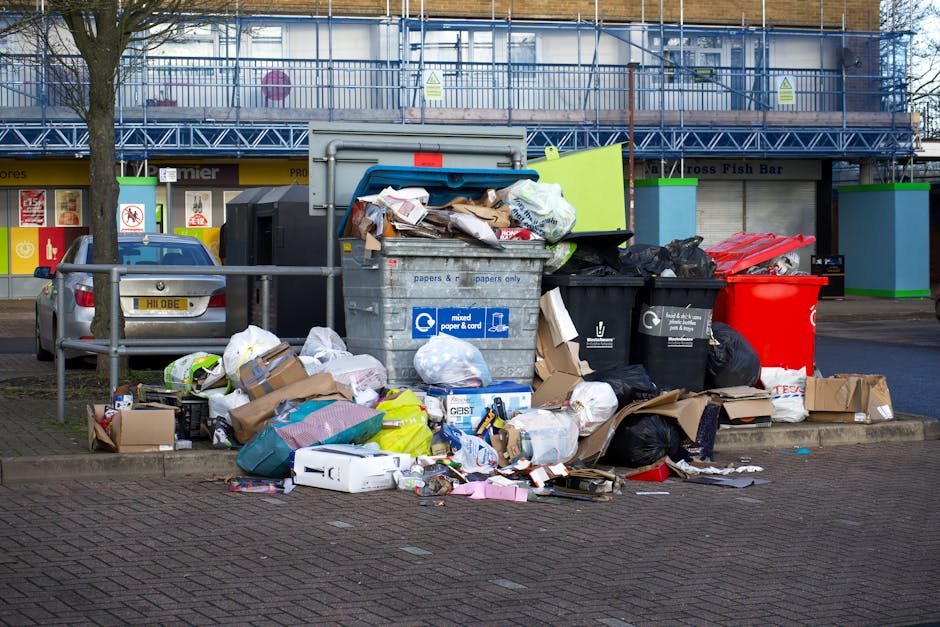 A collection of discarded household items and debris piled on a concrete pavement outside a building. In the foreground, there are wooden frames or pallet slats leaning against each other, with a smal